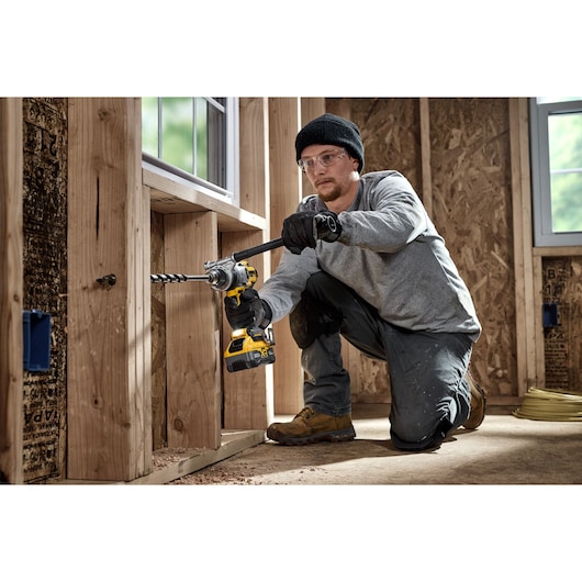 A person wearing work gloves and construction clothing is kneeling while using a DEWALT cordless drill to bore a hole in the wooden frame of a building under construction.