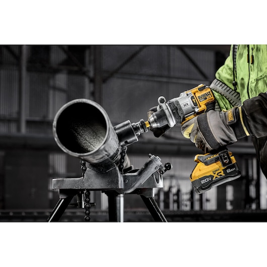 A person wearing safety gear is using a DEWALT cordless power drill to cut or drill into a large metal pipe, which is secured on a stand in an industrial setting.