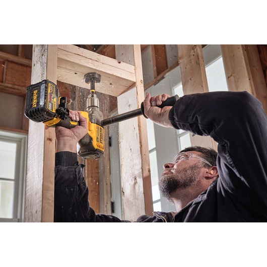 A person is using a DEWALT cordless drill with a hole saw attachment to drill through a wooden beam at a construction site. The person's face is blurred.