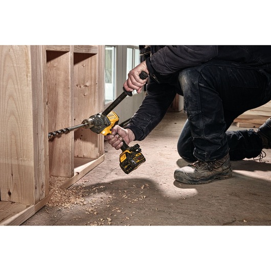 A person using a DEWALT cordless drill with an auger bit to drill a hole in a wooden structure, with wood shavings scattered on the floor.