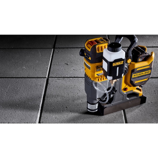 A DEWALT brushless cordless rotary hammer drill is shown positioned on a concrete tile floor. The tool features a water attachment and tubing for dust control or cooling. The product is primarily yellow and black with some white components.