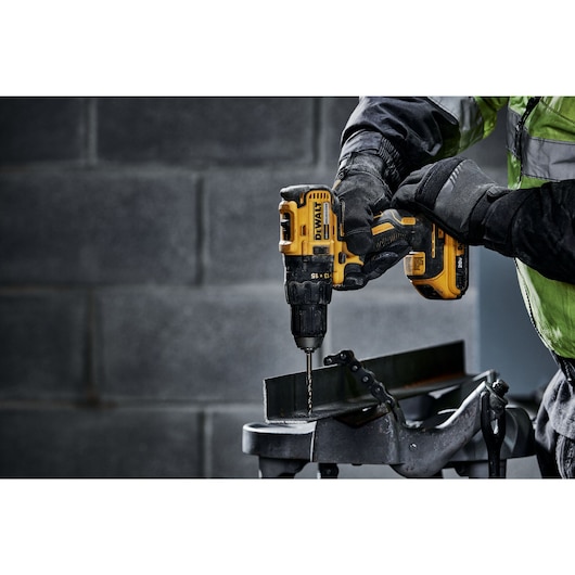 A person wearing protective gloves uses a yellow DEWALT cordless power drill to drill into a piece of metal that is held in a metal vise.