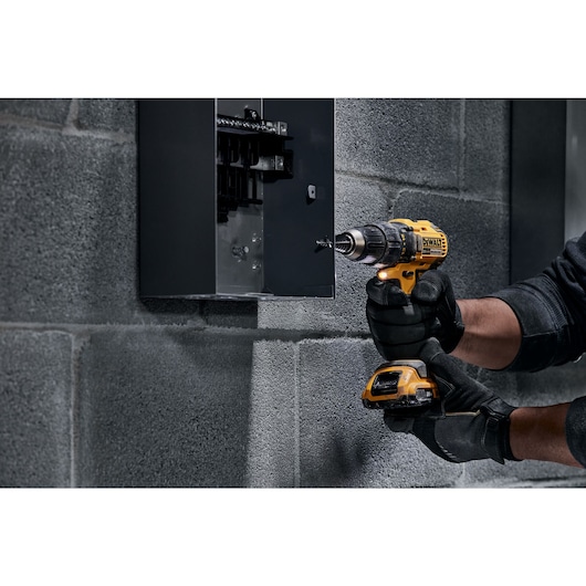 A person wearing black gloves is using a yellow DEWALT cordless drill to work on a gray electrical panel mounted on a concrete wall.