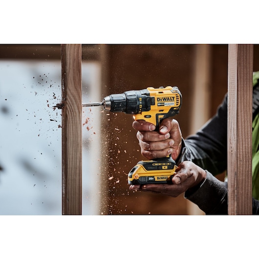 A close-up of a person using a yellow and black DEWALT cordless drill to drill into a wooden beam, with wood shavings flying from the hole.