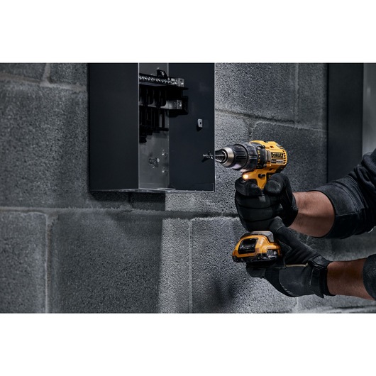 A person wearing black gloves is using a yellow DEWALT cordless drill to secure a screw into a gray electrical panel mounted on a concrete wall.
