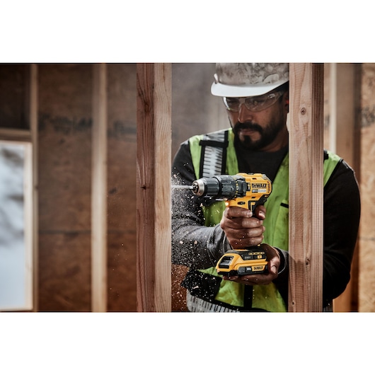 A person wearing a safety vest and helmet is using a yellow DEWALT cordless drill to drill into a wooden beam, creating wood shavings and dust.