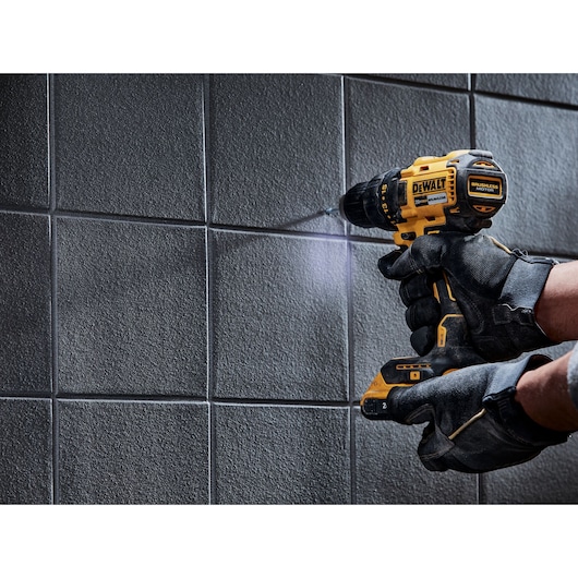 A person wearing black work gloves is using a yellow and black DEWALT cordless drill to drill into a tiled wall.