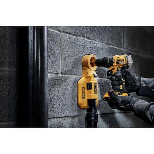 A person wearing gloves uses a yellow DEWALT cordless drill to drill into a concrete wall, with a DEWALT dust extraction attachment in place to collect dust.
