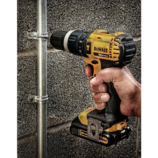 A hand holding a yellow and black DEWALT 20V Max cordless power drill, drilling into a metal pipe fastened to a concrete wall.