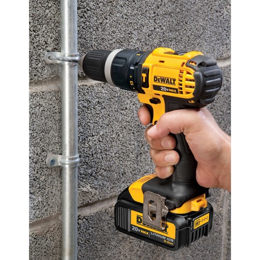 A hand holding a yellow and black DEWALT 20V MAX cordless drill, drilling a screw into a metal pipe clamp attached to a textured cement wall.