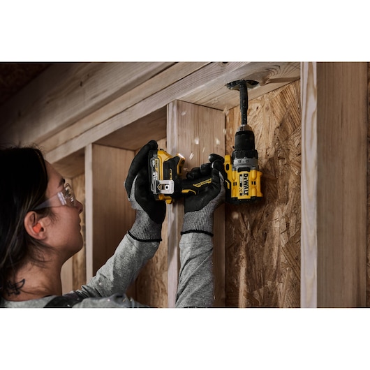 A person wearing work gloves is using a DEWALT cordless power drill to make a hole in a wooden construction frame or wall.