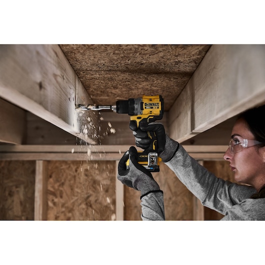 A person wearing gloves is using a DEWALT cordless drill to drill into a wooden beam, with wood shavings visibly falling.