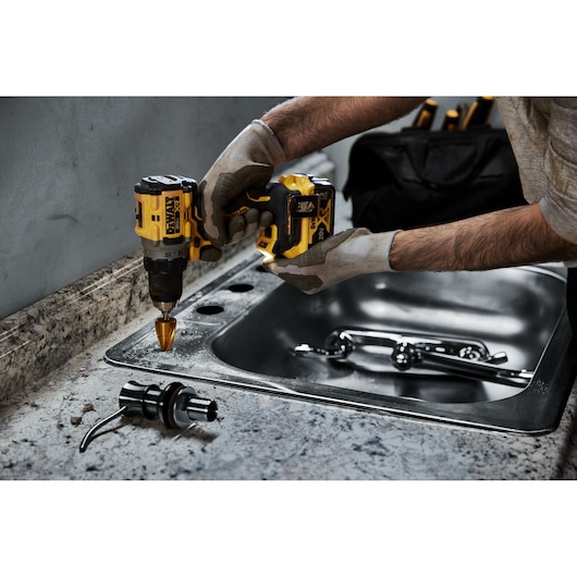 A person wearing gloves uses a yellow and black DEWALT cordless drill to drill holes into a countertop near a stainless steel sink. A faucet and plumbing part are visible on the counter. A black tool bag is in the background.