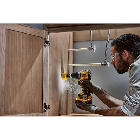 A person using a DEWALT cordless drill inside a wooden cabinet, drilling a hole near plumbing pipes. The person's face is blurred and they are wearing work gloves.