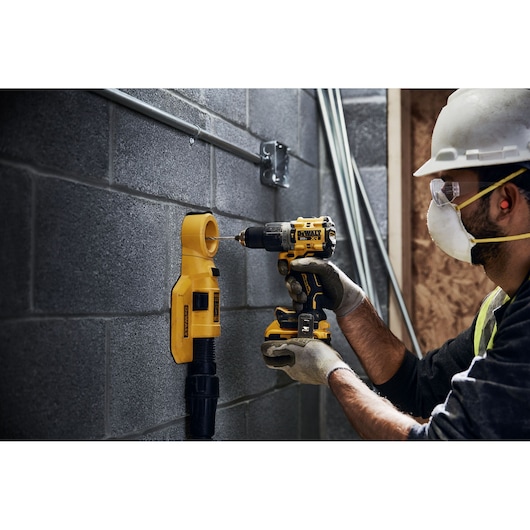 A person wearing safety gear uses a DEWALT DCD805B cordless drill on a concrete wall, with a DEWALT dust extraction attachment mounted below the drill site.