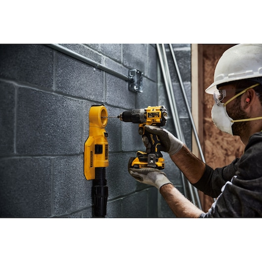 A person wearing work gloves and a hard hat is using a yellow and black DEWALT cordless drill to drill into a concrete block wall. A yellow DEWALT dust extractor attachment is positioned on the wall beneath the drill.