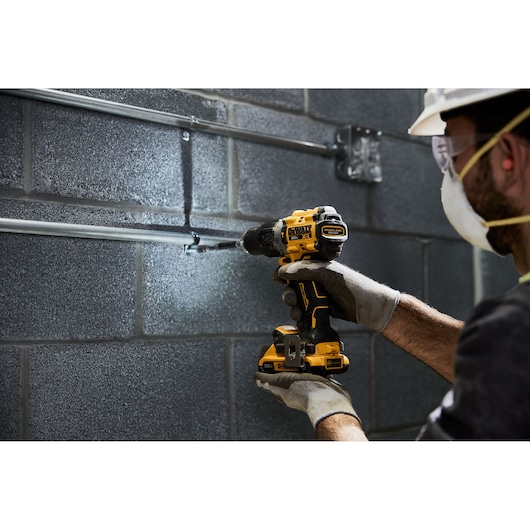A person wearing gloves and a safety helmet uses a yellow and black DEWALT cordless power drill to work on metal pipes attached to a gray cinderblock wall.