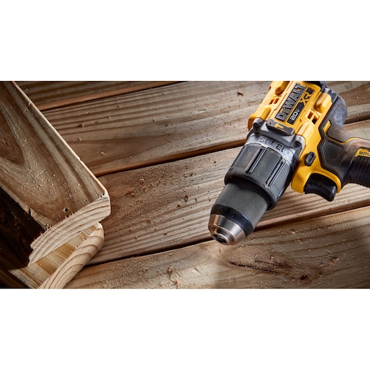 A close-up of a DEWALT cordless drill resting on a wooden surface with sawdust and a wood plank nearby.