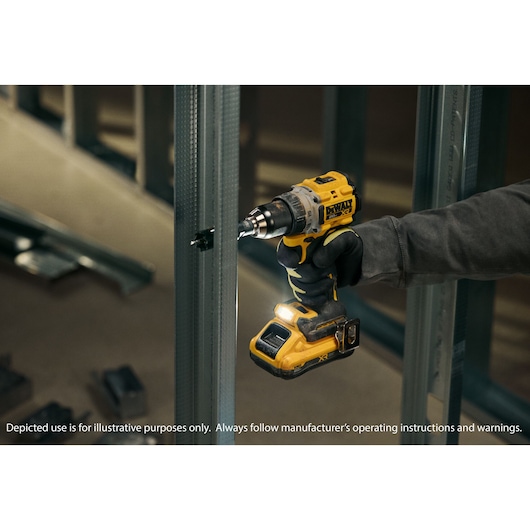 A person wearing gloves uses a yellow DEWALT cordless drill to screw into a metal stud frame in a construction environment. The drill features a built-in LED light. The setting is indoors, with construction materials and metal framing visible.