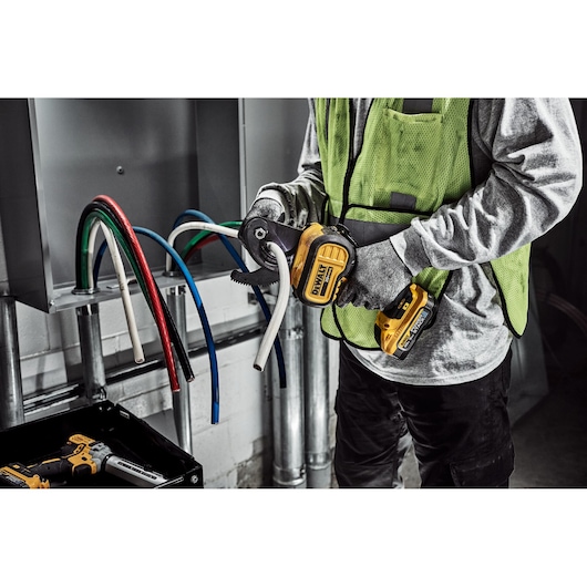 A person wearing gloves and a safety vest is using a DEWALT cordless cable cutter to cut a thick white electrical cable. Several colored cables are visible coming out of an electrical panel.