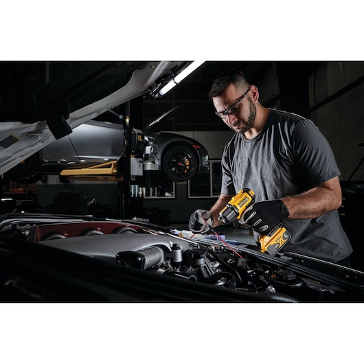 A person wearing gloves uses a DEWALT cordless heat gun (model DCE530B) to work on wiring under the hood of a vehicle in an automotive workshop.