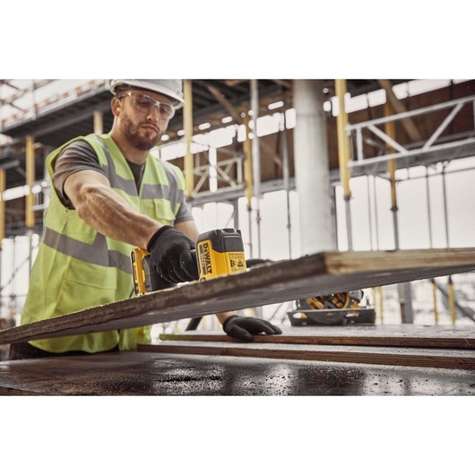 A construction worker wearing a safety vest and gloves is using a DEWALT DCE592D1 photography product on a wooden plank at a building site, with scaffolding visible in the background.