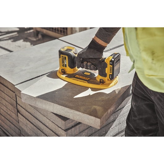 A person is using a DEWALT DCE592D1 cordless tool to lift a large concrete slab, with the tool's handle gripped firmly. The tool is shown on top of a stack of concrete slabs outdoors.