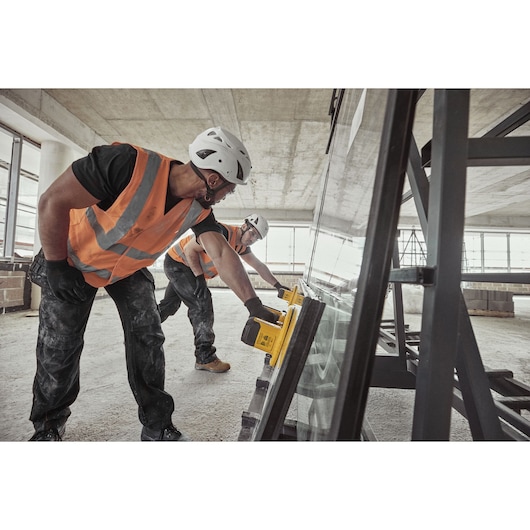Two workers in safety helmets and orange vests use a yellow DEWALT tool to assist with the installation or movement of a large sheet of glass in a construction site.