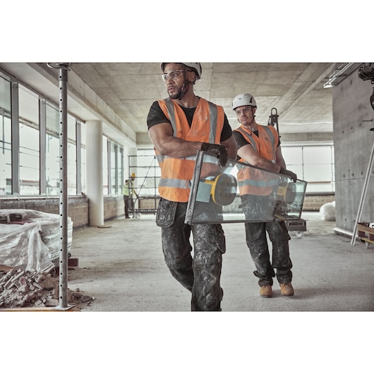 Two construction workers wearing orange safety vests and helmets are carrying a large pane of glass using DEWALT suction cups inside a building under construction.