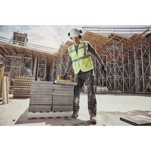 A person wearing safety gear and a high-visibility vest is using a DEWALT tool to lift a concrete slab at a construction site. Scaffolding and building materials are visible in the background.