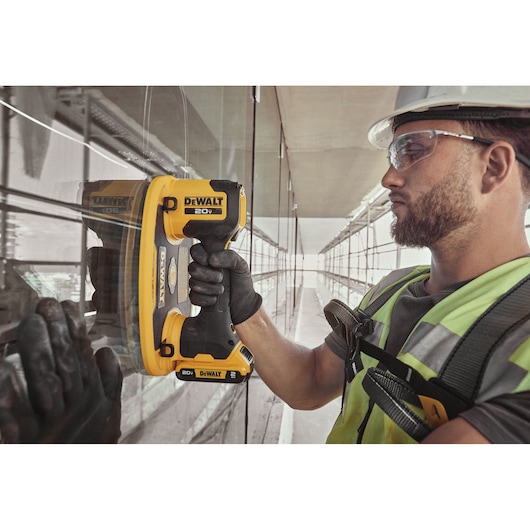 A person wearing safety gear uses a DEWALT 20V battery-powered suction cup tool to lift or install a glass panel in a construction or industrial setting. The person's face is blurred for privacy.