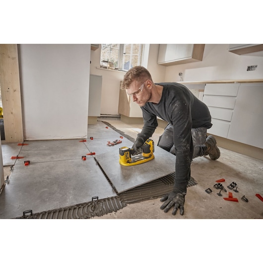 A person wearing work gloves is using a DEWALT DCE592D1 tool to install large ceramic tiles on a floor in a kitchen renovation setting. Tile spacers and adhesive are visible on the floor.