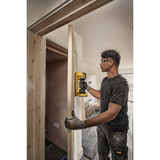 A person wearing work gloves is using a DEWALT power tool to sand or finish a wooden door frame in a construction or renovation setting. Their face is blurred for anonymity.