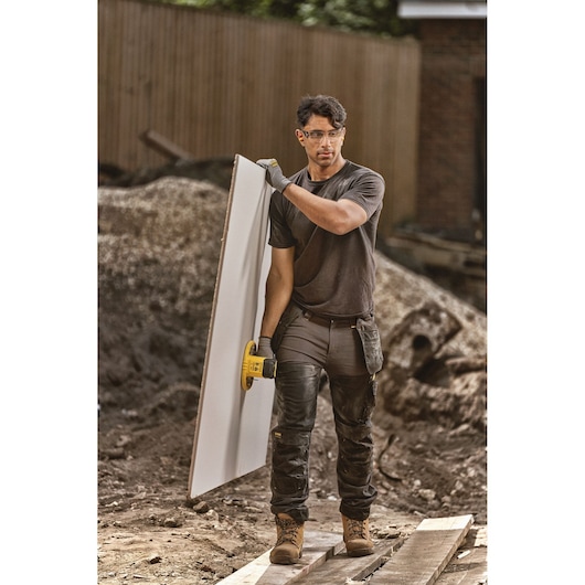 A person wearing work gloves and construction attire is carrying a large panel using a yellow DEWALT tool. The scene is an outdoor construction site with gravel, dirt, and wooden planks visible in the background.