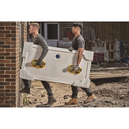 Two people wearing work gloves use DEWALT suction cup handles to carry a large white rectangular piece, possibly a shower tray, through a construction site doorway. The ground is uneven, and building materials are visible in the background.