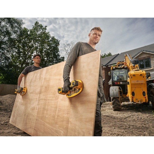 Two people carrying a large wooden board using DEWALT cordless suction tools at a construction site, with a yellow construction vehicle and a house under construction in the background.