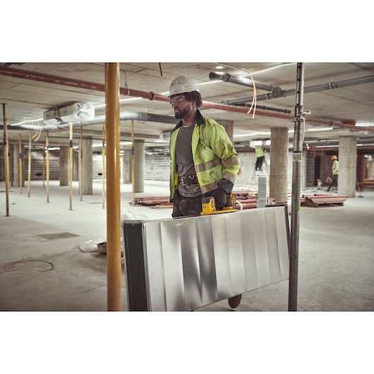A worker at a construction site is carrying a large metal panel using a DEWALT tool. The worker is wearing a yellow reflective jacket, gloves, and a white safety helmet. The background shows concrete pillars and other workers in safety gear.