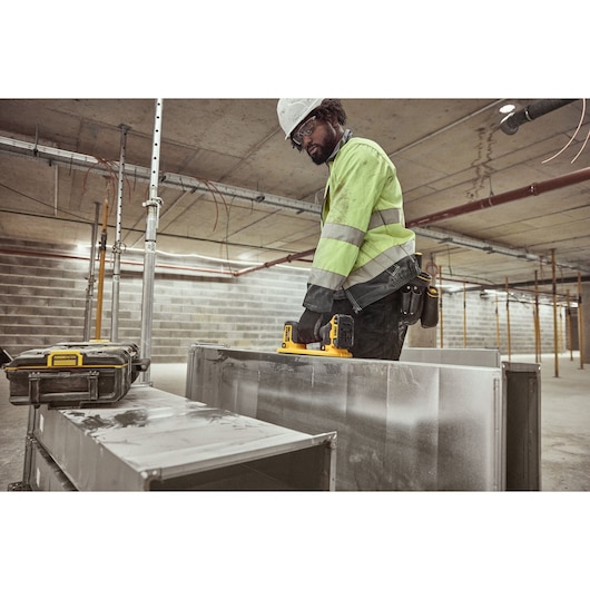 A worker wearing a safety helmet and high-visibility jacket is using a DEWALT portable work light (SKU: DCE592D1) on a metal duct in an unfinished industrial construction site. A DEWALT tool case is also visible nearby.