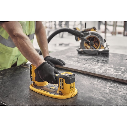 A person wearing black gloves uses a DEWALT cordless sander on a dark wooden surface, with a DEWALT circular saw visible in the background.