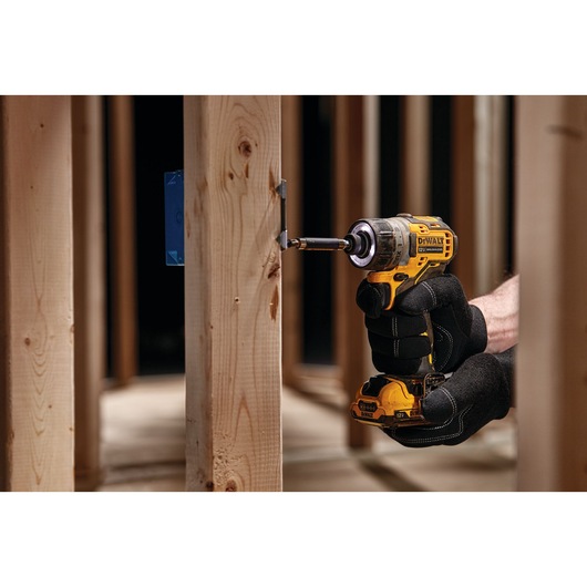 A person wearing black gloves uses a DEWALT cordless impact driver to drive a screw into a wooden stud frame at a construction site.
