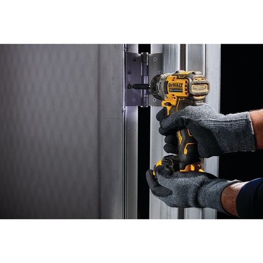 A person wearing grey work gloves uses a DEWALT cordless screwdriver to fasten a hinge on a metal door frame.
