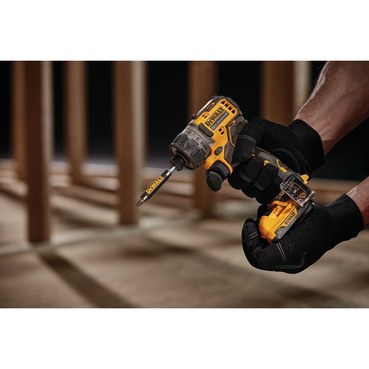 A person wearing black gloves is holding a yellow and black DEWALT cordless drill with a DEWALT drill bit attached. The background shows wooden beams and a construction setting.