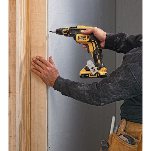 A person using a DEWALT DCF620D2 cordless screwdriver to fasten drywall to a wooden frame. The individual is wearing a tool belt and work clothes.