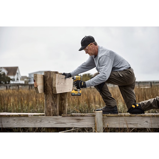 A person wearing work gloves and protective gear uses a DEWALT cordless drill to attach wooden boards to a large wooden post outdoors. The person is kneeling on a wooden platform near tall grass, with houses visible in the background.