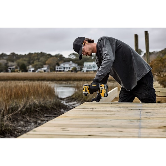 A person wearing a cap and gloves is using a DEWALT cordless drill to fasten wood planks outdoors, near a marsh area with houses visible in the background. The individual is working on a wooden deck or dock.