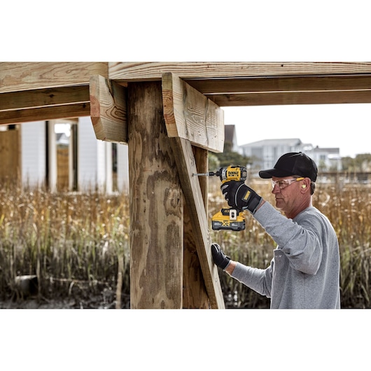Person wearing gloves and a cap using a DEWALT cordless impact driver to drill screws into outdoor wooden beams during construction.