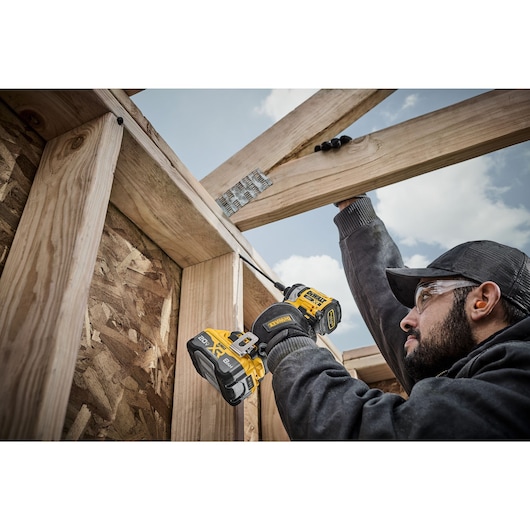 A person using a DEWALT DCF860B cordless drill to assemble wooden beams at a construction site. The individual's face is obscured, and the drill is visibly marked with the DEWALT logo.
