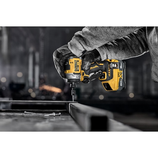 A person wearing gloves uses a DEWALT 20V cordless impact driver to fasten a screw into a metal frame in an industrial setting.