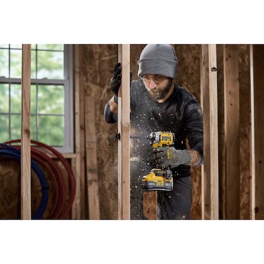 A person wearing gloves and a hat is using a DEWALT DCF860B cordless power tool to drill into a wooden frame in a construction setting. Sawdust is flying as the tool is operated.