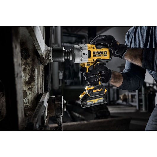 A person wearing gloves uses a DEWALT 20V XR cordless impact wrench to work on a large metal machine part in an industrial setting.